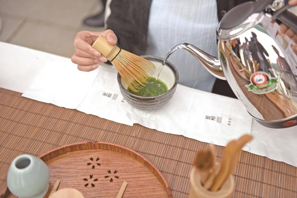 Ceremonial-grade matcha being whisked with a bamboo chasen in a small dark ceramic bowl, hot water poured from a steel kettle